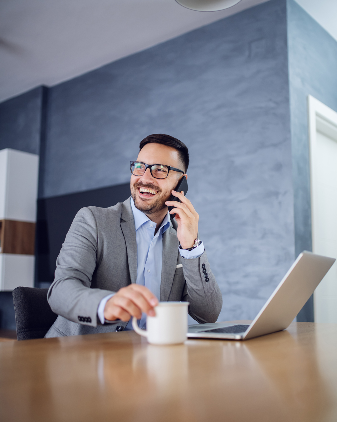 Man laughing on the phone while seated with laptop and mug